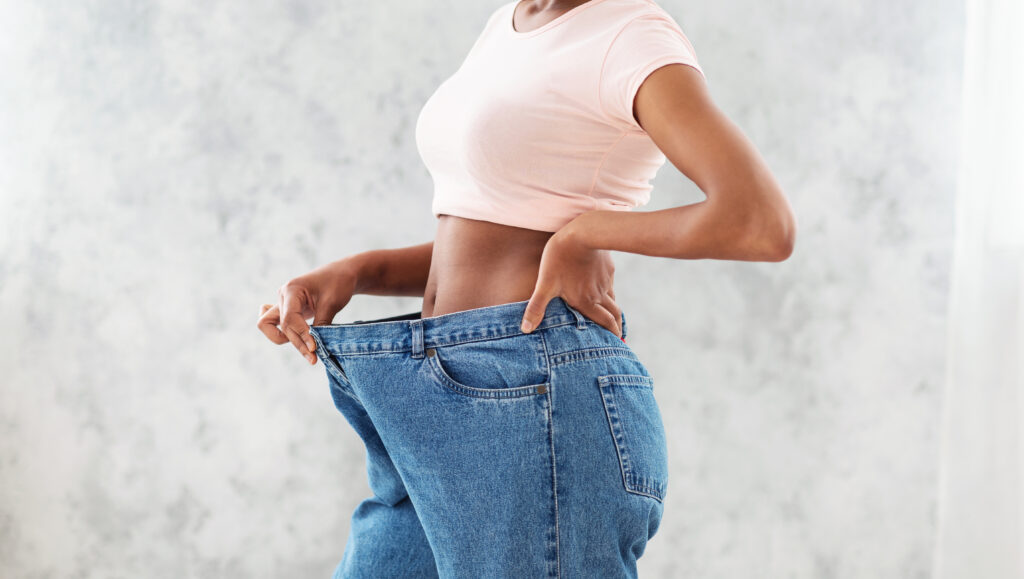 Unrecognizable black woman in oversized jeans showing results of her weight loss diet or liposuction, light background